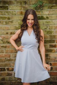 Smiling woman with long wavy brown hair in a light blue sleeveless dress stands by a weathered brick wall.