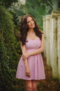 Woman with long brown hair in a pink dress smiles by a white stone wall, surrounded by green plants.