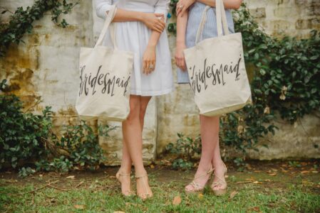 Legs of two women in light dresses with “Bridesmaid” tote bags, on grass by a vine-covered brick wall.