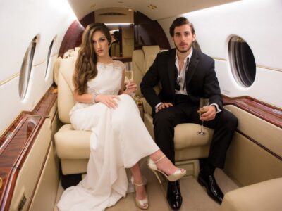 A woman in a white dress and a man in a black suit enjoy champagne on a private jet, facing the camera.