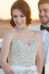 Bride in a beaded gown with choker smiles holding bouquet beside groom in white tuxedo at sunset.