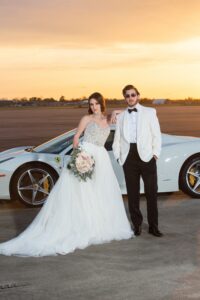 Bride in white gown with bouquet and groom in white tuxedo pose by white sports car at sunset on open pavement.