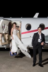 Woman in a white dress exits private jet as man in black tuxedo stands nearby at night.