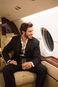 Man in black suit smiling with champagne in a cream leather seat on a private jet.