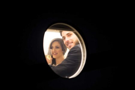 Smiling man and woman raise glasses, their reflection in a small round mirror against a dark background.