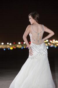 Woman in white beaded gown stands outdoors at night, facing away, with blurred city lights in the background.