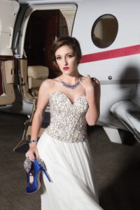 Woman in a strapless gown with blue jewelry holds blue heels by a private jet, looking at the camera.