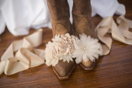 Brown cowboy boots with cream flowers and lace, surrounded by beige ribbon on a wooden floor.
