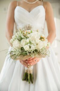 Bride in white dress holding a bouquet of white roses, babys breath, and greenery wrapped in burlap and lace.