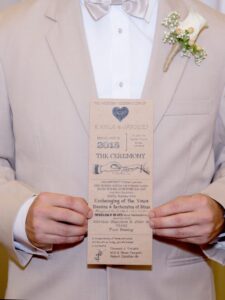 Man in beige suit and bow tie holds wedding program; white boutonnière with greenery on left lapel.