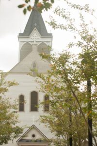 White church with arched windows and steeple partly hidden by green trees on a cloudy day.