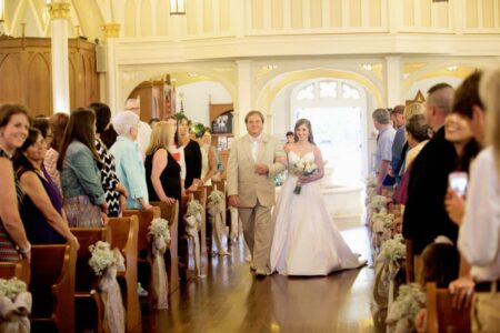 Bride in white walks down aisle with older man in tan suit, surrounded by smiling guests in a decorated church.