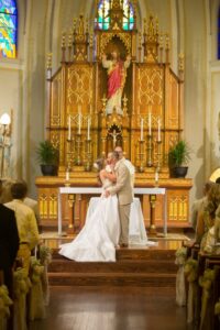 Bride and groom kiss at the church altar during their wedding, with guests watching and stained glass in the background.