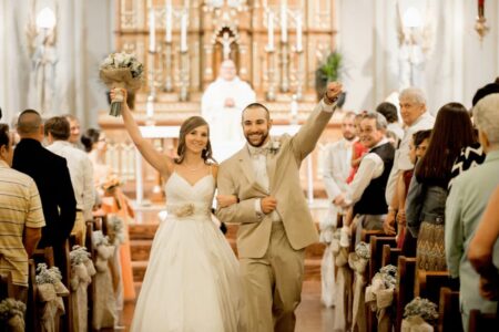 Bride and groom walk down the aisle holding hands, celebrating as guests cheer in a decorated church.