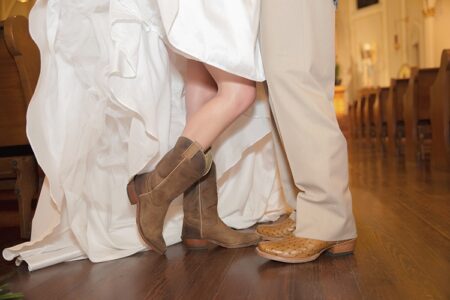 Bride in white dress and cowboy boots stands with groom in light pants and woven shoes on a wooden floor.