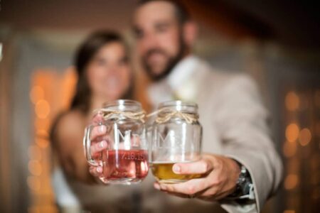 Bride and groom toast with mason jar drinks labeled Mr. and Mrs., smiling with a warm, softly lit background.