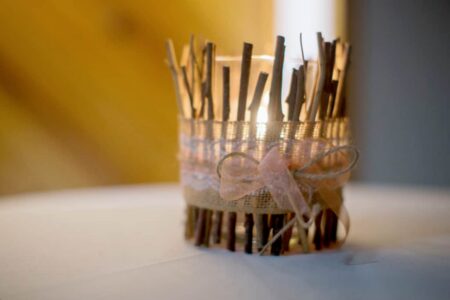 Lit candle in a glass holder with twigs, burlap, lace, and ribbon bow on white surface, blurred background.