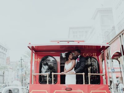Bride and groom in wedding attire smiling on a red trolley with city buildings and palm trees in the background.