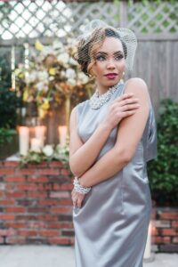 Woman in silver satin dress with pearl jewelry, birdcage veil, and styled hair poses by brick wall with flowers and candles.