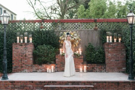 Woman in white gown on brick patio with candles, greenery, floral backdrop, and two tall lampposts.
