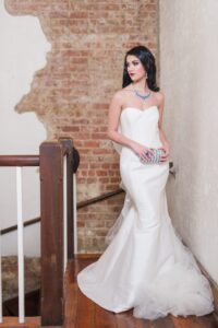 Bride in a strapless white gown with blue jewelry holds a clutch on wooden stairs by a brick wall, looking to the side.