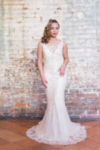 Bride in sleeveless white gown and birdcage veil poses by rustic brick wall, with curled hair over one shoulder.