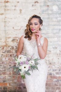 Bride in lace dress with birdcage veil holds white and lavender bouquet by brick wall, one hand touching her face.