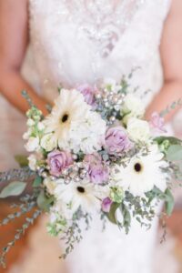 Bride holding a bouquet of white gerbera daisies, pale pink roses, greenery, and small white flowers with a blurred background.