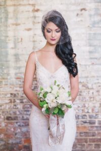 Bride in beaded white dress holds white rose bouquet, standing by rustic brick wall with birdcage veil over long dark hair.
