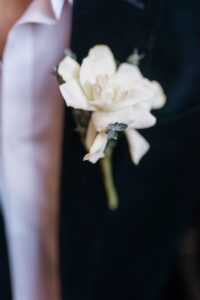 White rose boutonniere pinned to a dark suit lapel, with a white dress shirt in the background.