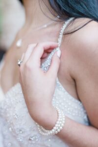 Woman in beaded dress and pearl jewelry touches her shoulder strap, long dark hair flowing, wearing a delicate necklace.
