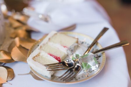White cake with red filling on a decorative plate, two forks, gem-shaped objects, and gold leaves on a white tablecloth.