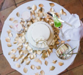 White cake with a white flower on top sits on a table with gold leaves, utensils, drinks, and a sliced piece, shot from above.