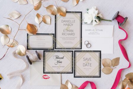 Elegant wedding stationery flat lay with invites, RSVP, thank you note, rings, gold leaves, white flower & pink ribbon.