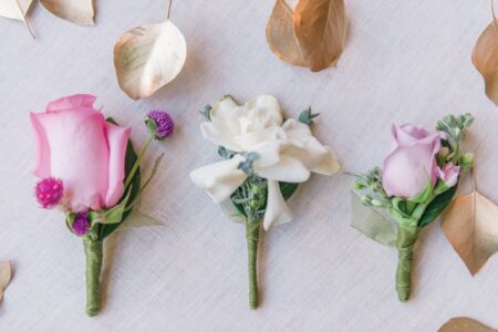 Three boutonnières—pink rose, white flowers, and lavender rose—rest on light fabric with scattered gold leaves.