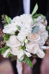 Bouquet with white roses, greenery, and accent flowers held by someone in a black suit.