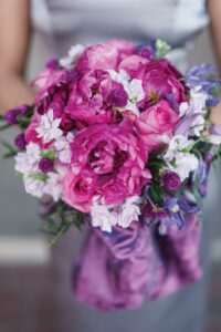 Person in gray outfit holding bouquet of pink peonies, purple flowers, and white blossoms wrapped in purple fabric.