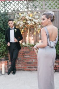 Woman in silver gown with birdcage veil holds clutch; man in tuxedo leans on brick wall with flowers and candles.