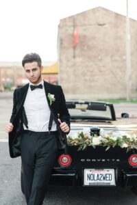 Man in tuxedo stands by black Corvette with flowers and NOLA2WED plate, outside a brick building.