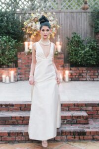 Woman in cream gown and pearls poses gracefully on brick steps amid candles and greenery outdoors.