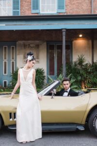 Woman in white dress stands by vintage gold convertible; man in tuxedo sits inside. Blue-shuttered building, tropical plants behind.