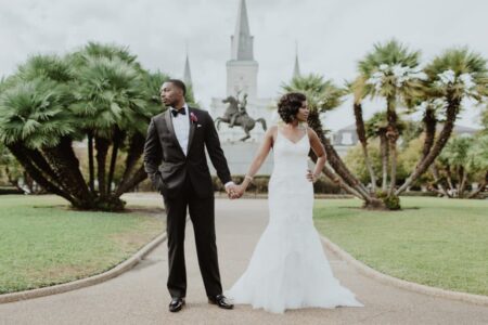 Bride and groom hold hands in a park, facing away; he wears a tuxedo, she a white gown. Palm trees and statue behind them.