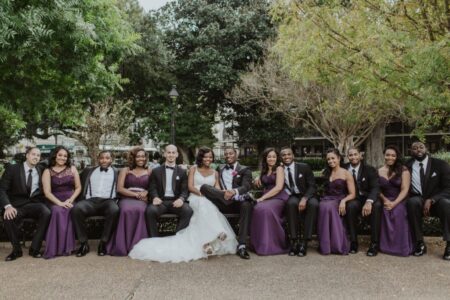 Bride, groom, and wedding party in purple dresses and black suits smile outdoors amid trees and greenery.