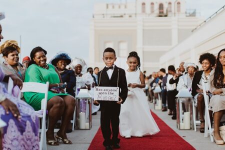 A boy in a suit carries a “Has anyone seen the rings?” sign on the red carpet, followed by a girl in white, as guests smile.