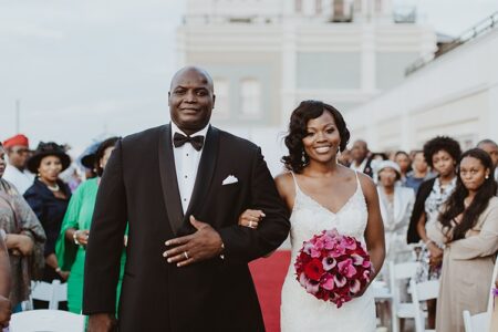 Bride in white dress and man in tuxedo walk down red aisle at outdoor wedding, surrounded by guests.