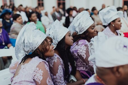 Women in lavender lace dresses and silver headwraps sit together at an event, with others seated attentively in the background.