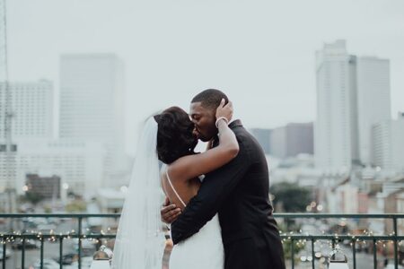 Bride and groom kiss on a balcony with city buildings behind them; she wears a white dress and veil, he a black suit.
