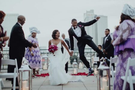 Bride and groom walk down an outdoor aisle; groom jumps joyfully. Guests smile amid rose petals with city buildings behind.