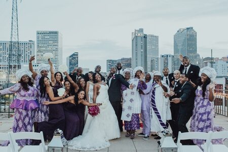 Wedding party in purple and white poses on a rooftop with city skyline, bride and groom smiling at the center.