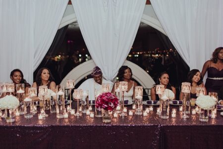 Seven people dine at a candlelit table with white flowers, elegant decor, and city lights visible through large draped windows.